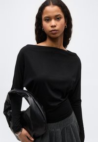 Νεαρή γυναίκα με μακριά, σγουρά μαλλιά φοράει μαύρο μπλουζάκι με λαιμόκοψη βάρκα και γκρι φούστα με λεπτές ρίγες, κρατώντας μια μαύρη δερμάτινη τσάντα στο αριστερό της χέρι.