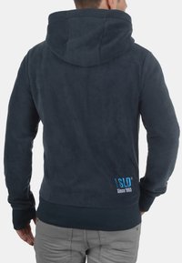 Sudadera con capucha de forro polar azul marino con una textura suave, un bolsillo frontal y un detalle de logo en azul claro en la parte baja de la espalda. Incluye un cordón en la capucha.