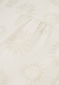Weißer Stoff mit einem subtilen Glanz, der ein sich wiederholendes Sonnenmotiv in hellem Gold zeigt. Die Textur ist glatt und hat gefaltete Details.