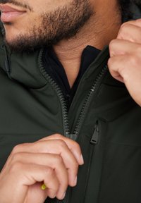 Grønne zip-jakker med høj krave, tekstureret stof og kontrasterende sort indvendig foer. Lynlåsdetaljer er synlige. Hånd justerer kraven.