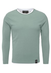 T-shirt a maniche lunghe in cotone verde chiaro con colletto tondo bianco e un'etichetta nera con il marchio nella parte inferiore sinistra. Texture liscia.