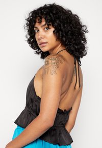 Top peplum preto em seda com decote halter, costas abertas e acabamento com franzidos. Modelo exibe uma tatuagem floral no ombro. Saia azul visível.