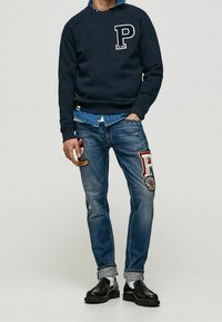 Sweat-shirt navy avec logo "P" blanc, accompagné d'un jean bleu usé avec des patches colorés et de mocassins noirs.