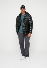 Chaqueta acolchada negra con capucha, que presenta una textura brillante y costuras horizontales. Llevada sobre una camisa azul, combinada con pantalones grises y zapatos blancos.