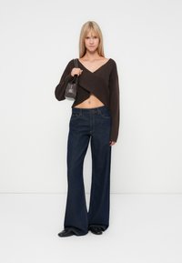 Un pull court marron avec un design enveloppant, associé à un jean bleu foncé à taille haute et des chaussures noires. Le modèle tient un petit sac à main gris.