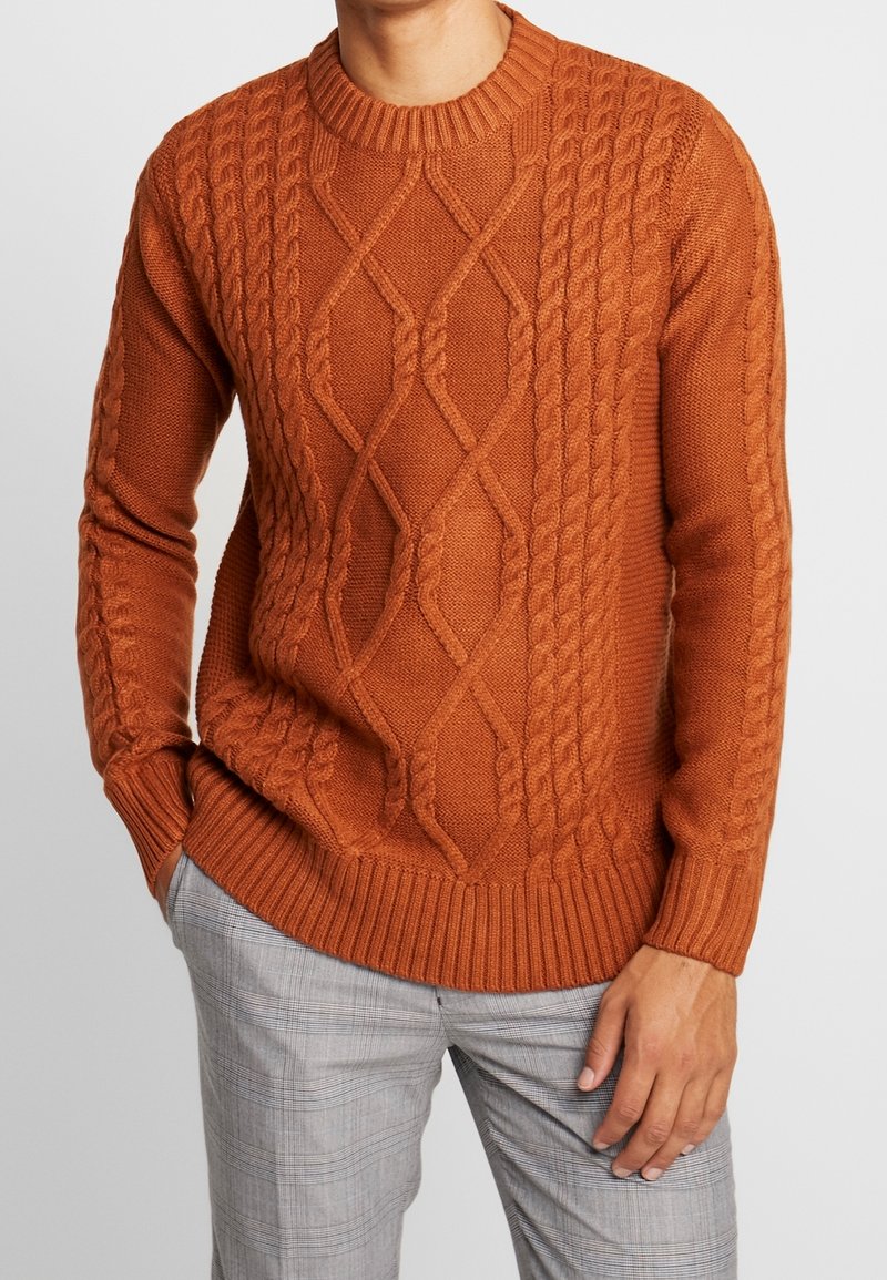 Homme portant un pull à torsades orange rouille et un pantalon à carreaux gris clair, debout une main dans la poche sur un fond uni.