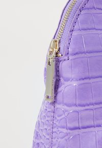 Lila torbica iz umetne krokodilske kože s sijajno površino, opremljena z zlatim zadrgo in majhno oznako z blagovno znamko "Ted Baker."