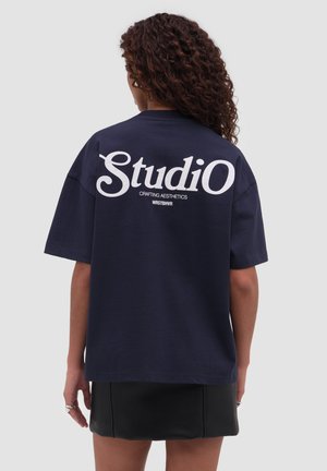 Kobieta o kręconych włosach, ubrana w granatowy oversize'owy T-shirt z napisem "Studio Crafting Aesthetics WRSTBHVR" na plecach oraz czarną spódnicę.
