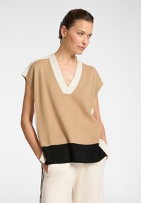 Top sin mangas de punto en color beige con escote en V, detalles en crema en los hombros y un dobladillo negro. Presenta un corte holgado y bolsillos laterales.
