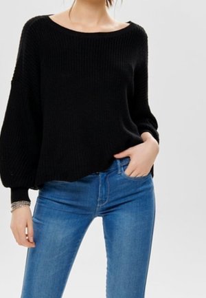 Femme portant un pull noir ample avec des manches bouffantes et un jean bleu ajusté, une main dans la poche, sur fond clair uni.