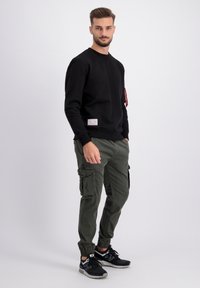 Sudadera negra con cuello redondo y etiqueta lateral, combinada con pantalones cargo verdes oliva que cuentan con bolsillos laterales y puños elásticos. Zapatillas deportivas negras.