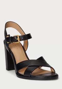 Čierne kožené sandále na vysokom opätku so širokým designom krížových remienkov, uzáverom na pracku okolo členka a pevným blokovým opätkom. Hladká matná textúra.