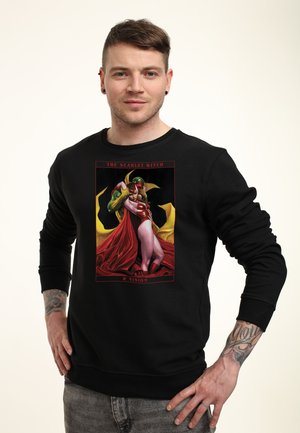 Mężczyzna w czarnej bluzie z kolorową grafiką Scarlet Witch i Vision z przodu, stoi z rękami na biodrach.