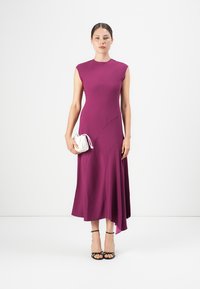 Donna in piedi che indossa un vestito midi senza maniche di colore viola, mentre tiene una piccola borsa bianca, abbinata a tacchi neri con cinturini.