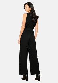 Črna brezrokavna kombinezon s prilegajočim pasom in širokimi hlačnicami, izdelan iz gladkega materiala. Ima detajl pasu in visok ovratnik.