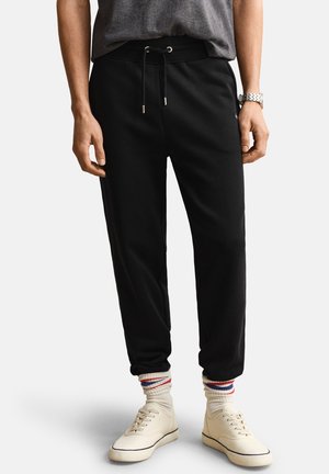Parte inferiore del corpo di una persona che indossa pantaloni da jogging neri con coulisse, scarpe da ginnastica bianche, calzini bianchi con strisce rosse e blu e una maglietta grigia.