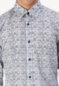 Lichtblauw knoopshirt met complexe donkerblauwe geometrische patronen, voorzien van een kraag en zwarte knopen aan de voorkant.
