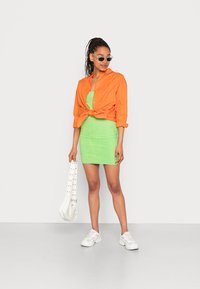 Camisa curta laranja abotoada e atada à cintura, combinada com um vestido justinho verde. Acessórios incluem ténis brancos e uma bolsa branca.
