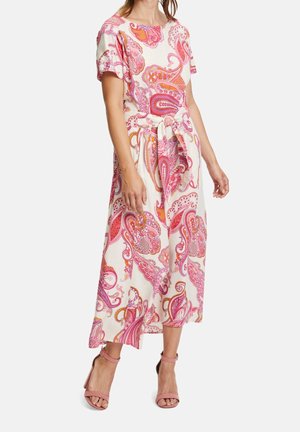 Kobieta ubrana w białą sukienkę midi z różowo-pomarańczowym nadrukiem w stylu paisley oraz wiązaniem w talii z przodu, zestawioną z różowymi szpilkami na kostkę.