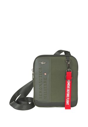 Borsa a tracolla quadrata verde oliva con tracolla nera, emblema argentato e portachiavi rosso con la scritta "REMOVE BEFORE FLIGHT".