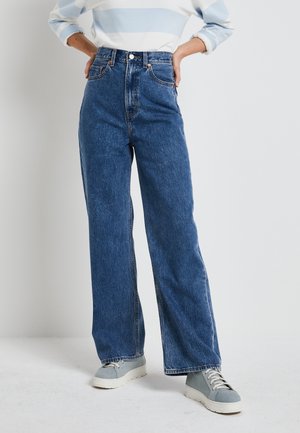 Personne portant un jean large taille haute bleu, un haut à manches longues rayé bleu clair et blanc, et des baskets bleu clair, debout avec les mains sur les hanches.