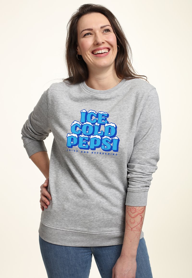 Felpa grigia con maniche lunghe, decorata con la scritta blu "ICE COLD PEPSI" e motivi di neve. I jeans in denim sono visibili, con un tatuaggio a motivi sul braccio.