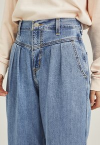 Korkeavyötäröiset, vaaleansiniset farkut, joissa on laskokset edessä, napitus, vyölenkit ja-taskut. Sileä pinta.