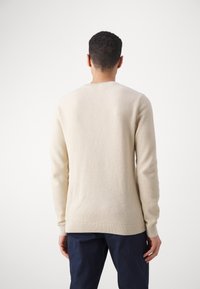 Beige stickad tröja med en strukturerad yta, rund hals och ribbade muddarna. Bärs med marinblå byxor, vilket visar en enkel, klassisk design.