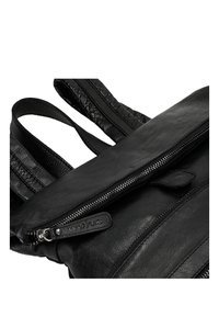 Uno zaino in pelle nera presenta un compartimento principale con zip, una superficie liscia e spallacci densi con finitura trapuntata. Etichetta del marchio visibile.
