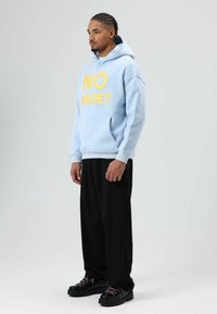 Sweat à capuche bleu clair avec texte jaune "NO MADE ?" sur le devant, poche kangourou, associé à un pantalon large noir et des chaussures noires avec des lacets roses.