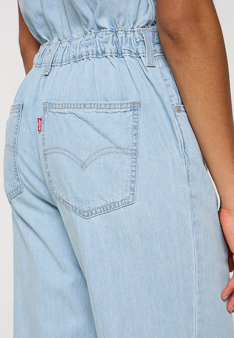 Jumpsuit en denim bleu clair doté d'une taille élastique, de deux poches arrière avec des coutures emblématiques et d'une coupe décontractée.