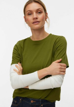 Olive green cotton t-shirt with short sleeves, layered over a white ribbed long sleeve top. The model is crossing her arms, wearing dark jeans.