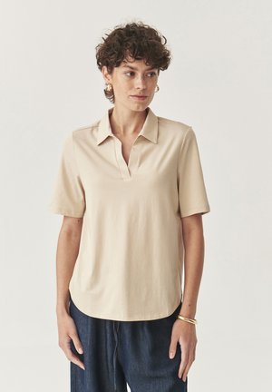 Polo shirt beige a maniche corte con scollatura a V, realizzato in tessuto morbido. Presenta una vestibilità rilassata e un design del colletto discreto. Indossato con pantaloni blu scuro.