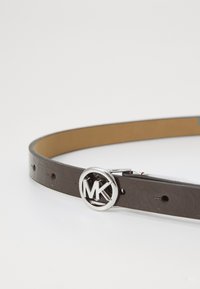 Cintura in pelle marrone con una fibbia circolare argentata lucida che presenta le lettere "MK". La cintura ha delle perforazioni per le regolazioni della misura.
