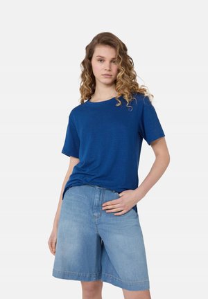 Giovane donna con capelli ricci che indossa una maglietta blu a maniche corte e pantaloncini di jeans chiari fino al ginocchio, in piedi con una mano sui fianchi.