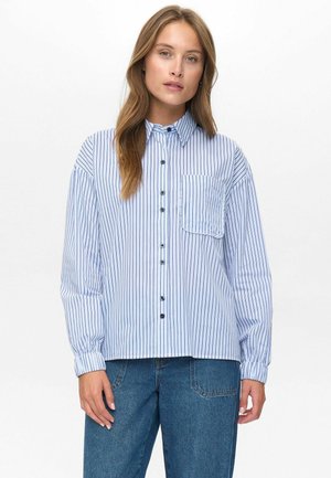 Woman wearing a blue and white vertically striped button-up shirt with a chest pocket and blue jeans, standing against a plain background.