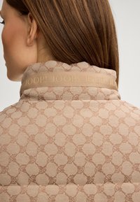 Eine Frau mit glattem braunem Haar, die eine beigefarbene gemusterte Jacke mit dem Text "JOOP!" auf dem Kragen trägt, von hinten und leicht zur Seite gezeigt.