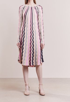 Femme portant une robe mi-longue à manches longues avec des rayures ondulées verticales en rose, violet, beige et marine, portée avec des talons couleur nude.