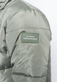 Ljusgrön puffjacka med en vadderad textur och en distinkt grön etikett med vit text som läser "LA ROSA COPENHAGEN."
