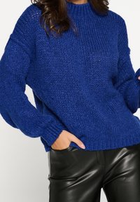 Pull en maille bleu épais avec un col montant, des manches larges et des poignets côtelés, associé à un pantalon en cuir noir, présentant une coupe décontractée.