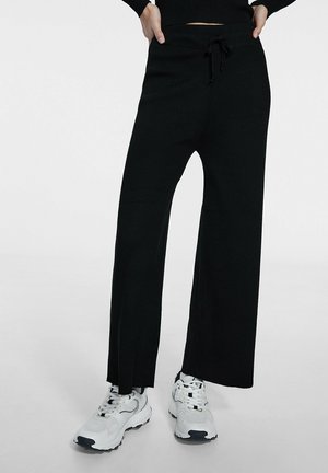 Persoon draagt zwarte, wijde broek, witte sportschoenen en een zwart shirt met lange mouwen, handen in de zakken, staand tegen een effen achtergrond.
