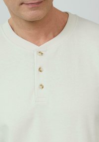 Helle beige Henley-Bluse mit V-Ausschnitt, drei Knöpfen, geripptem Kragen und strukturiertem Stoff. Schlichtes Design mit einer weichen, glatten Oberfläche.