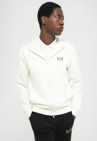 Haut de hoodie blanc à zip avec capuche et col court, arborant un logo EA7. Porté avec un pantalon noir, mettant en avant un design athlétique décontracté.