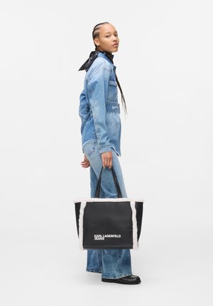 Nainen, joka pukeutuu farkkutakkiin ja farkkuihin, pitää mustaa kangaskassia, jossa on valkoinen eläinkarvatrimmi ja edessä "KARL LAGERFELD JEANS" -logo.