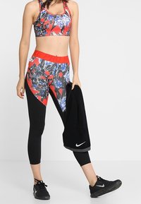 Top corto sportivo rosso con motivo floreale; leggings neri con accenti rossi; scarpe Nike nere classiche; tiene un asciugamano nero.