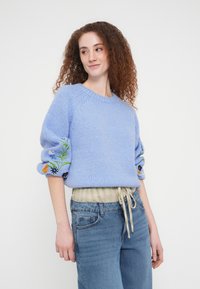Pull en tricot bleu clair avec manches bouffantes ornées de broderies florales colorées, associé à une chemise à carreaux et un jean en denim décontracté.