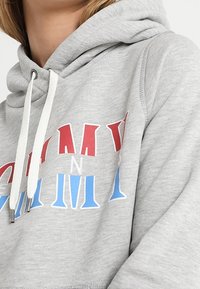 Zbliżenie na osobę w jasnoszarym kapturze z białymi sznurkami i kieszenią z czerwono-biało-niebieskimi literami w formacie blokowym.