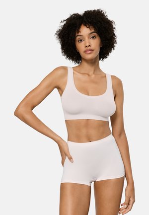 Frau mit lockigem Haar, die einen hellrosa Sport-BH und passende hoch taillierte Boyshorts trägt und vor einem schlichten weißen Hintergrund steht.