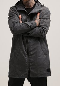 Long manteau gris avec fermeture éclair, deux poches avant, une capuche et un tissu texturé. Présente un accent de label noir au bas.