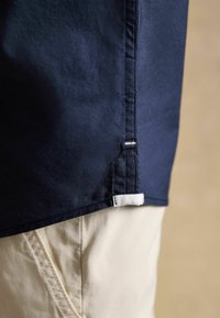 Gros plan sur l'ourlet d'une chemise bleu marine avec des détails de couture latérale portée sur un pantalon beige.